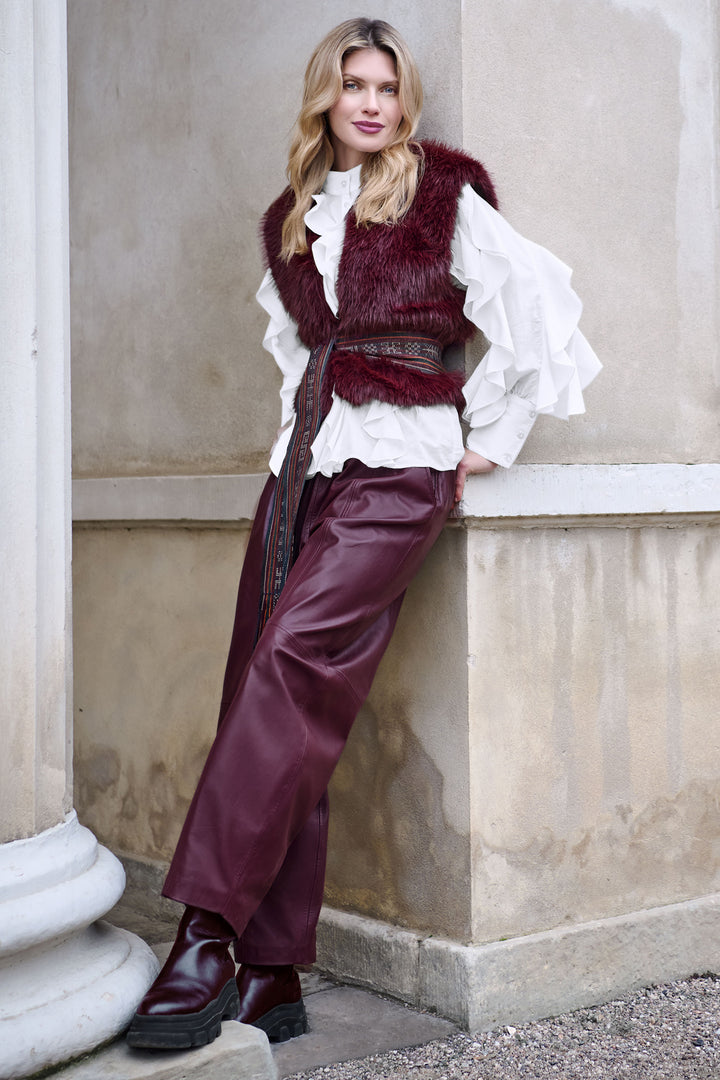 Woman in burgundy fur vest and pants leaning against a stone wall.