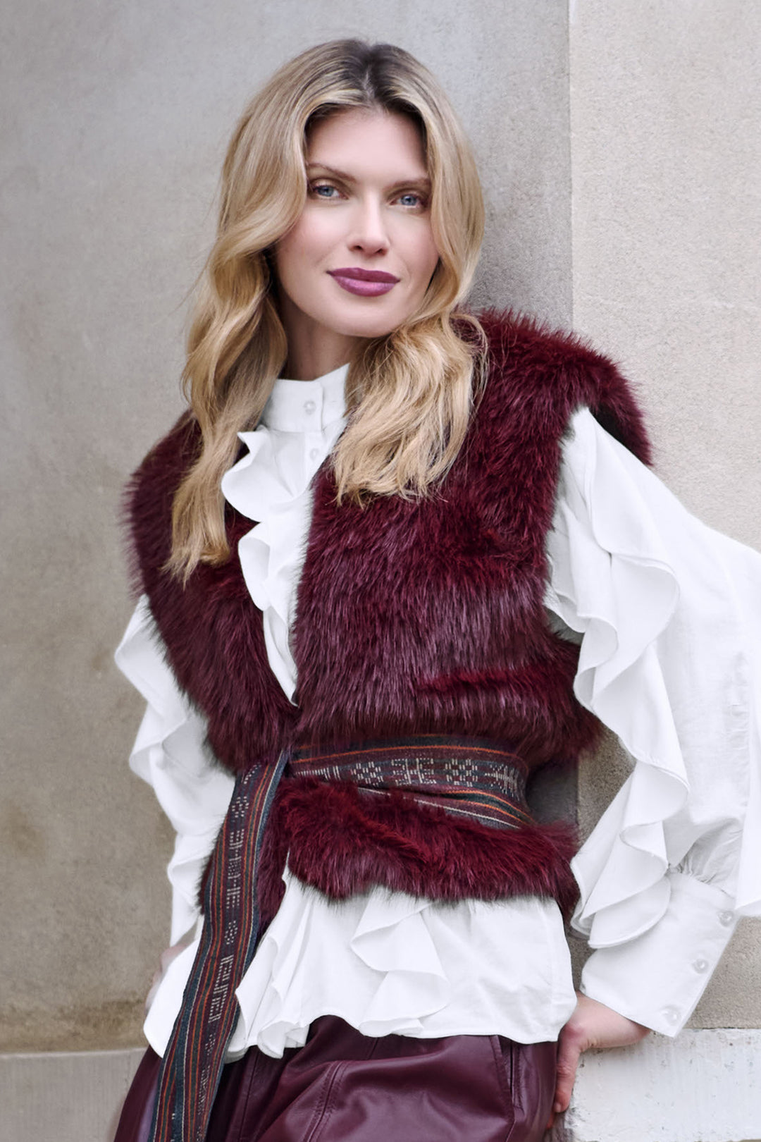 Woman wearing a burgundy fur vest over a white blouse against a neutral background