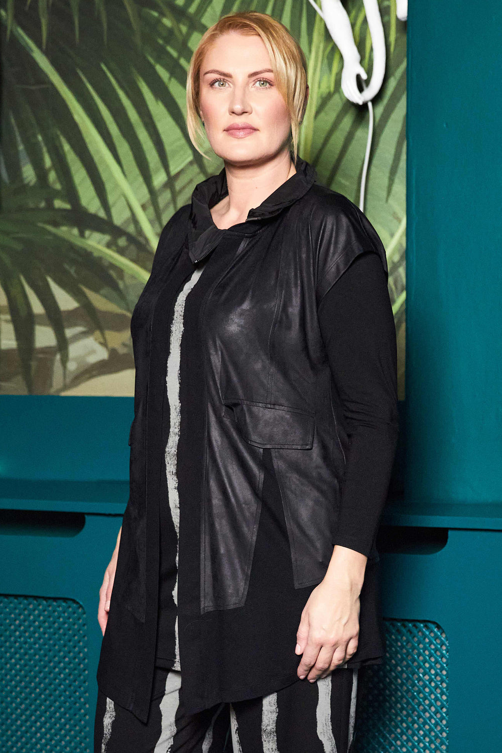 Person wearing a black leather jacket against a teal wall with green foliage.