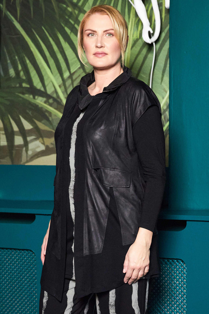Person wearing a black leather jacket against a teal wall with green foliage.