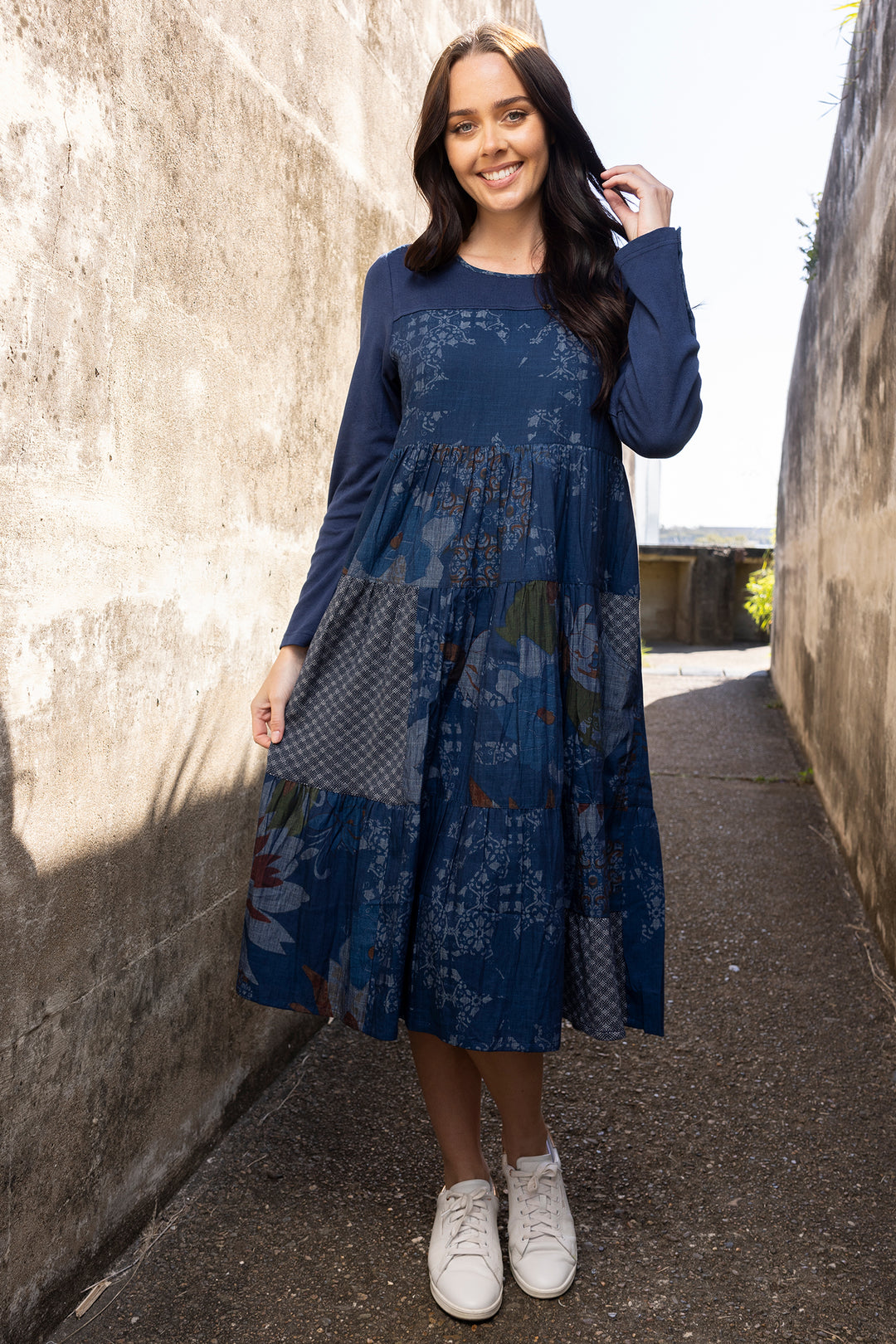 Woman wearing a blue floral dress standing in a sunlit alleyway.