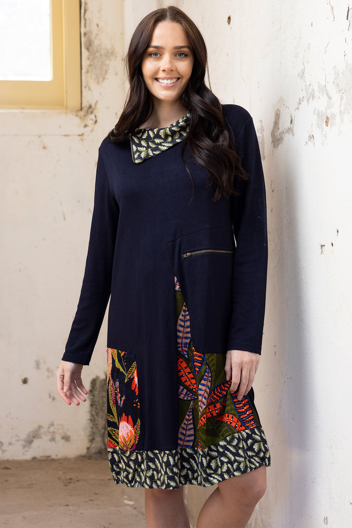 Woman wearing a navy dress with colorful patterns against a textured wall.
