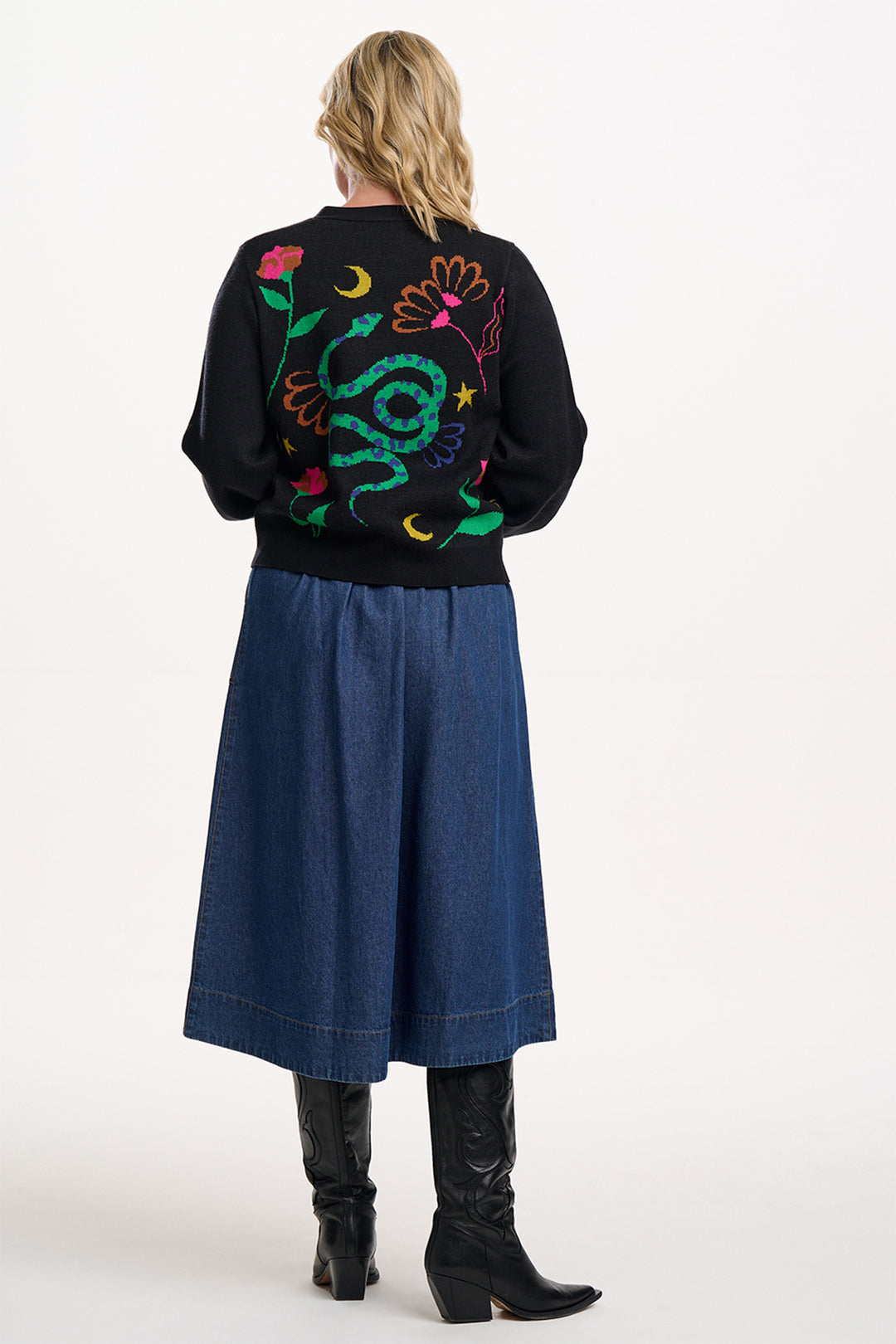 Person wearing a black sweater with colorful floral designs on the back, paired with a blue skirt and black boots.