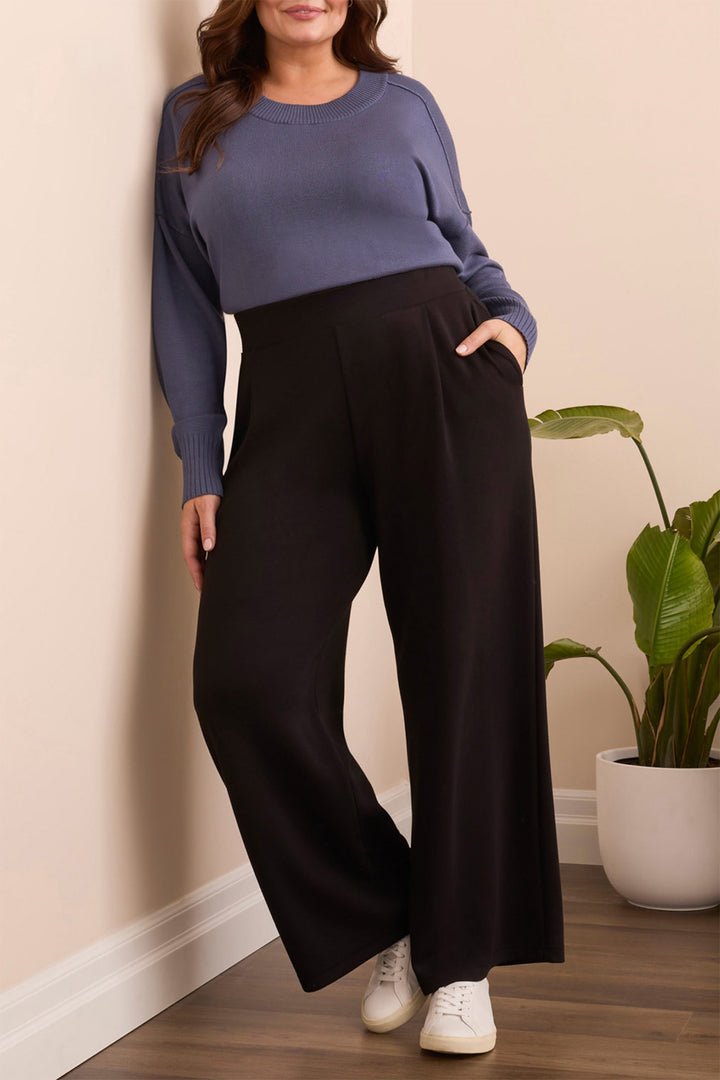 Woman wearing a blue sweater and black pants standing against a beige wall with a plant in the background.