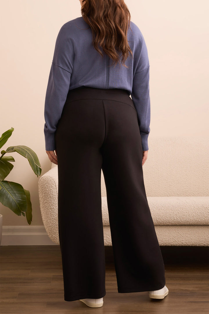 Person wearing a blue top and black pants standing in a room with a plant and sofa.
