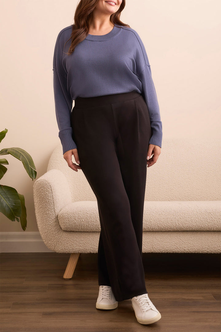 Woman wearing a blue sweater and black pants standing in a room with a beige couch and plant.