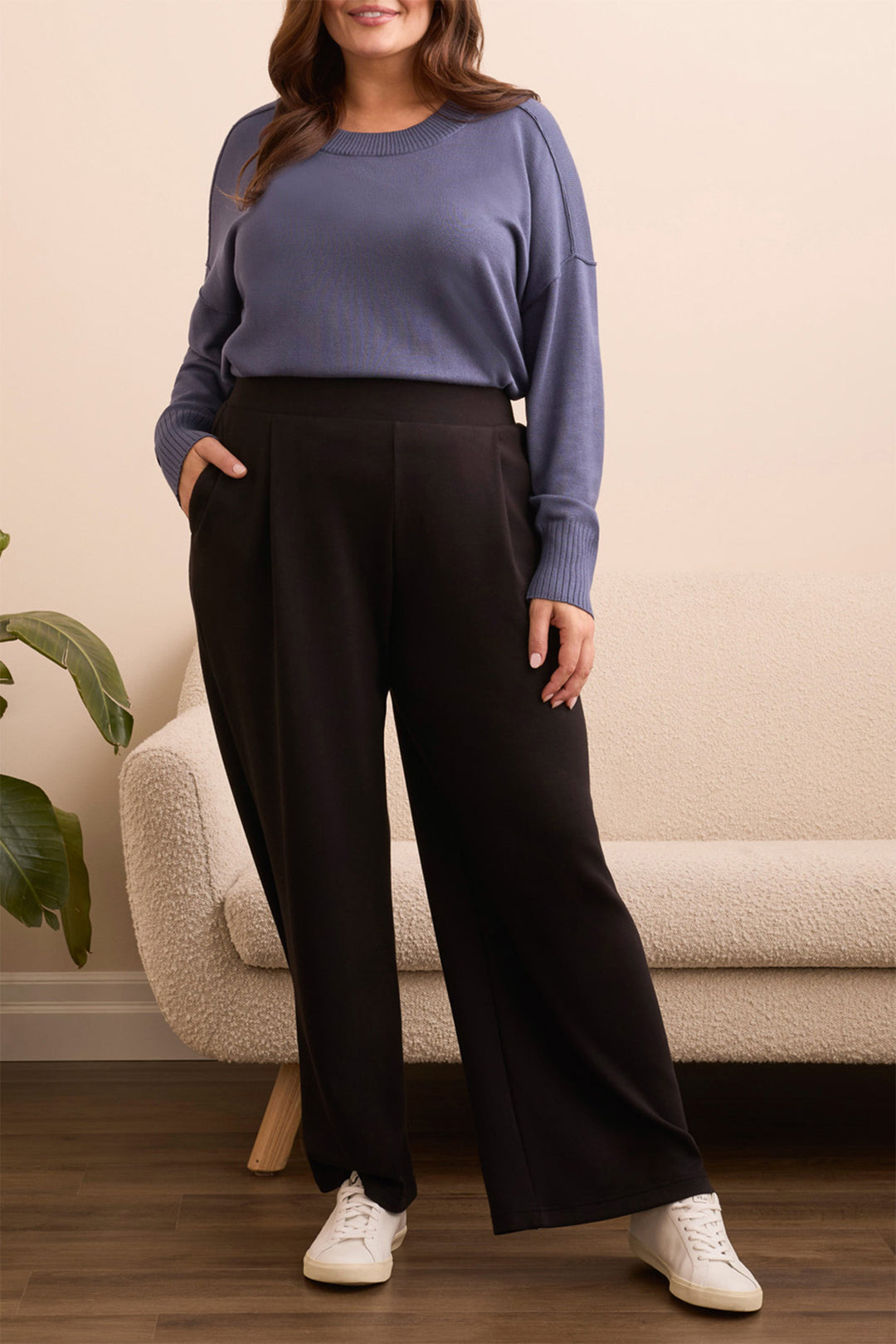 Woman wearing a blue sweater and black pants standing in a room with a beige sofa and plant.