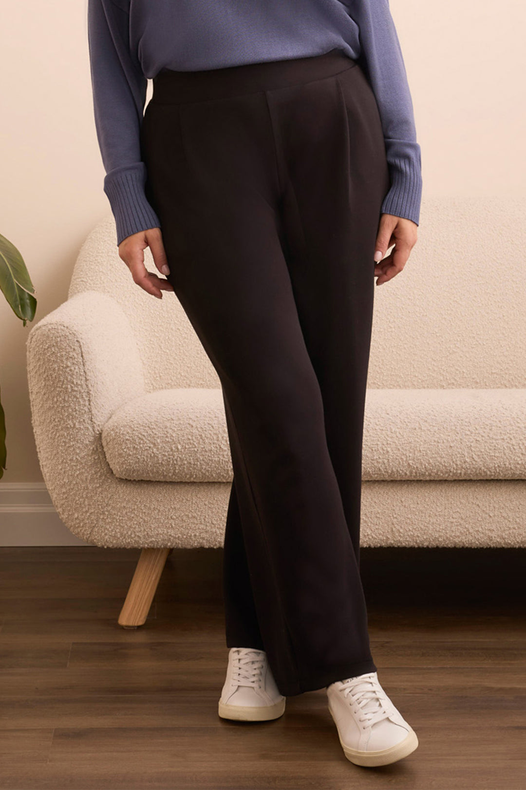 Person wearing black pants and a blue sweater standing in a room with a beige couch and wooden floor.