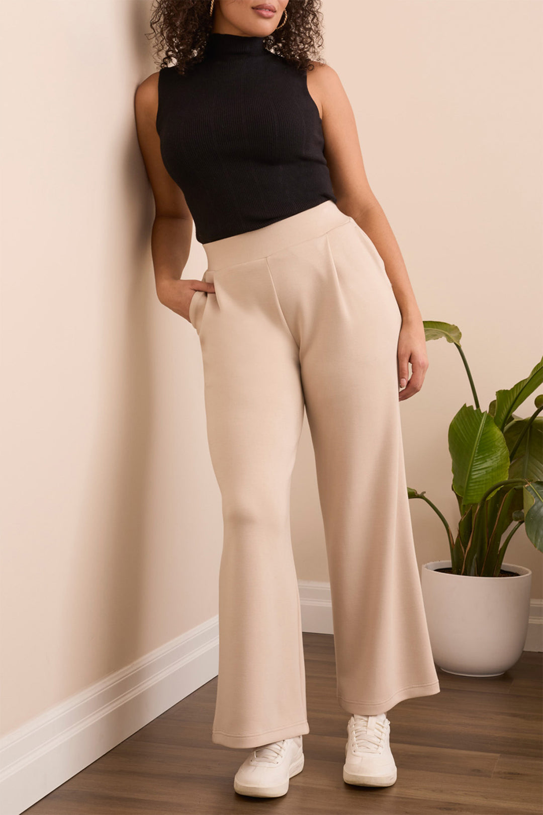 Woman wearing a black sleeveless top and beige wide-leg pants standing against a beige wall with a plant in the background.