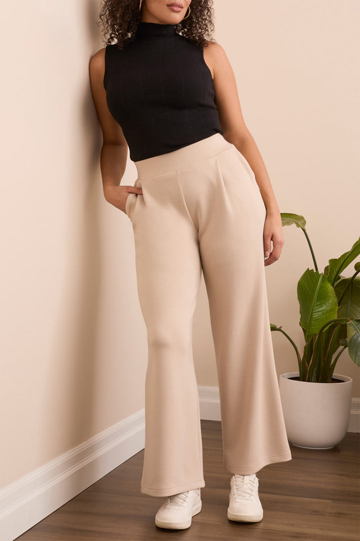 Woman wearing a black sleeveless top and beige wide-leg pants standing against a beige wall with a plant in the background.