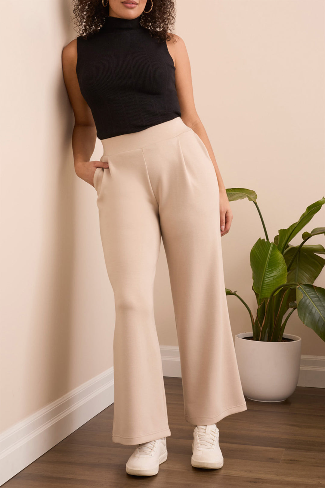 Woman wearing a black sleeveless top and beige wide-leg pants standing against a beige wall with a plant in the background.