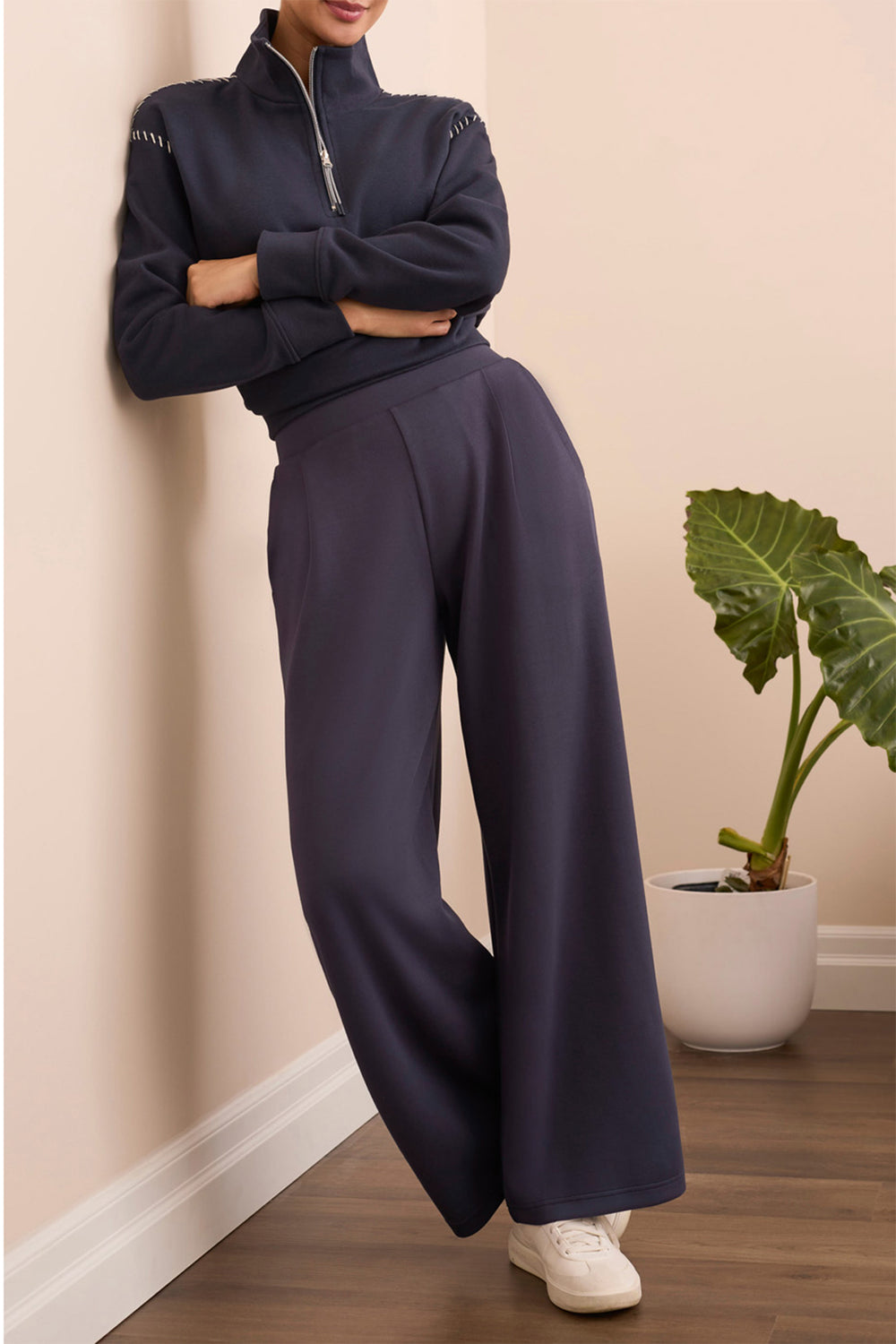 Person wearing a navy tracksuit standing against a beige wall with a plant in the background.