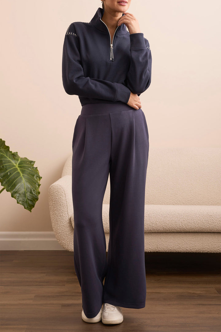 Person wearing a navy tracksuit standing in a room with a beige couch and green plant.