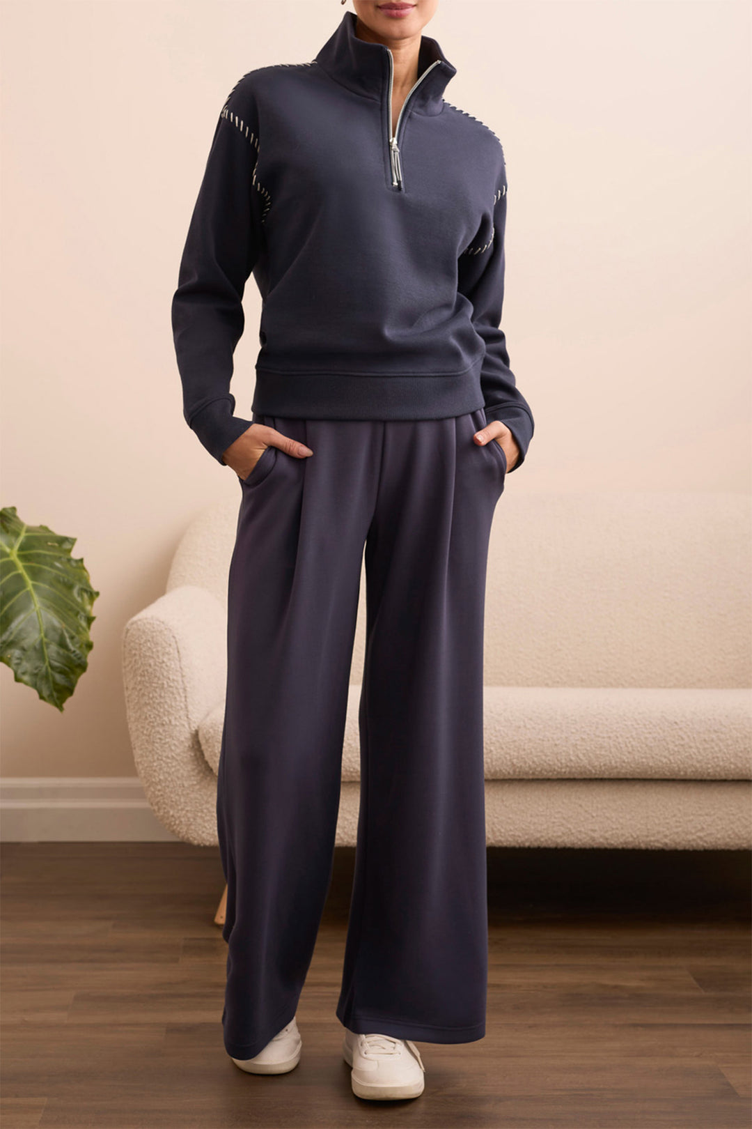 Person wearing a navy tracksuit standing in a room with a beige sofa and plant.