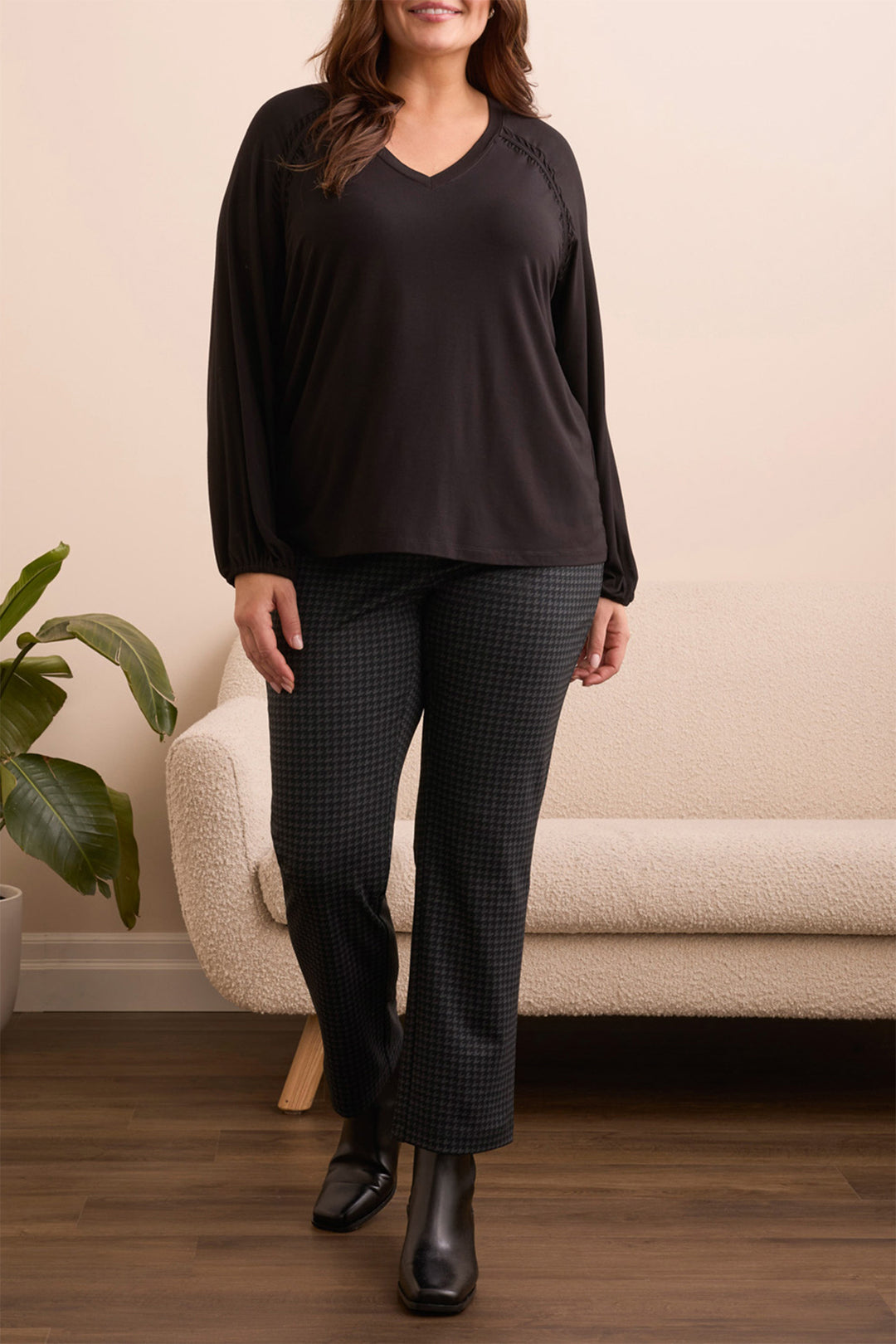 Woman wearing a black top and checkered pants standing in a room with a beige couch and plant.