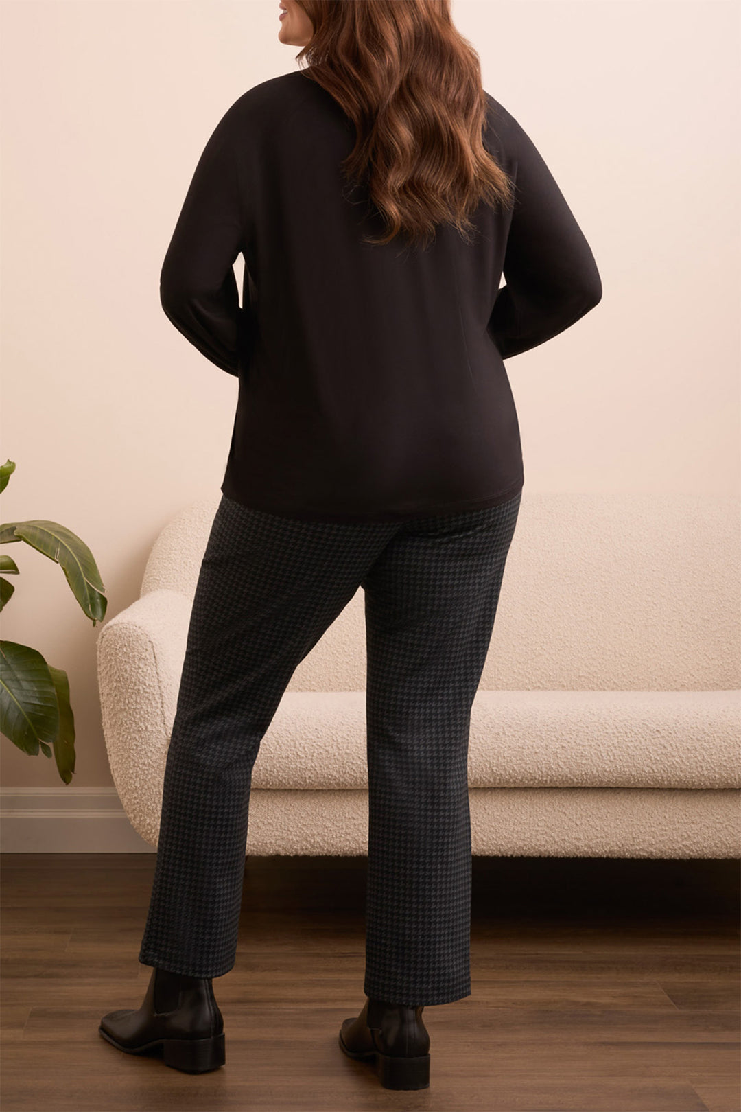 Person wearing a black sweater and plaid pants standing in a room with a beige sofa and plant.