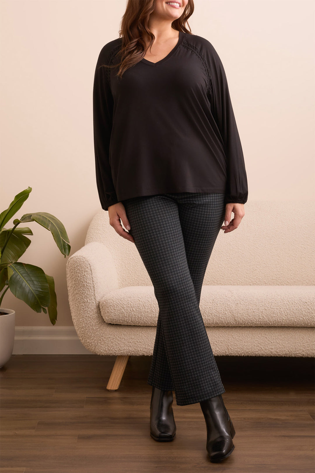 Woman wearing a black top and dark pants standing in a room with a beige armchair and plant.