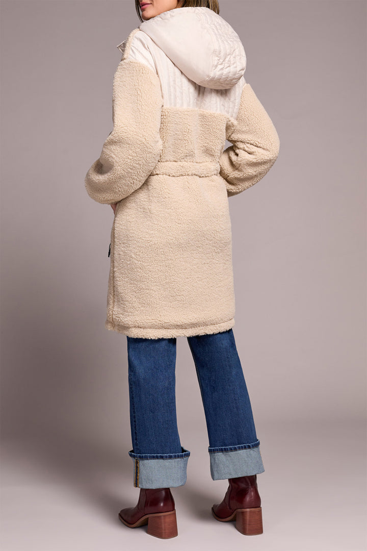 Person wearing a beige coat, white sweater, blue jeans, and brown shoes on a plain background