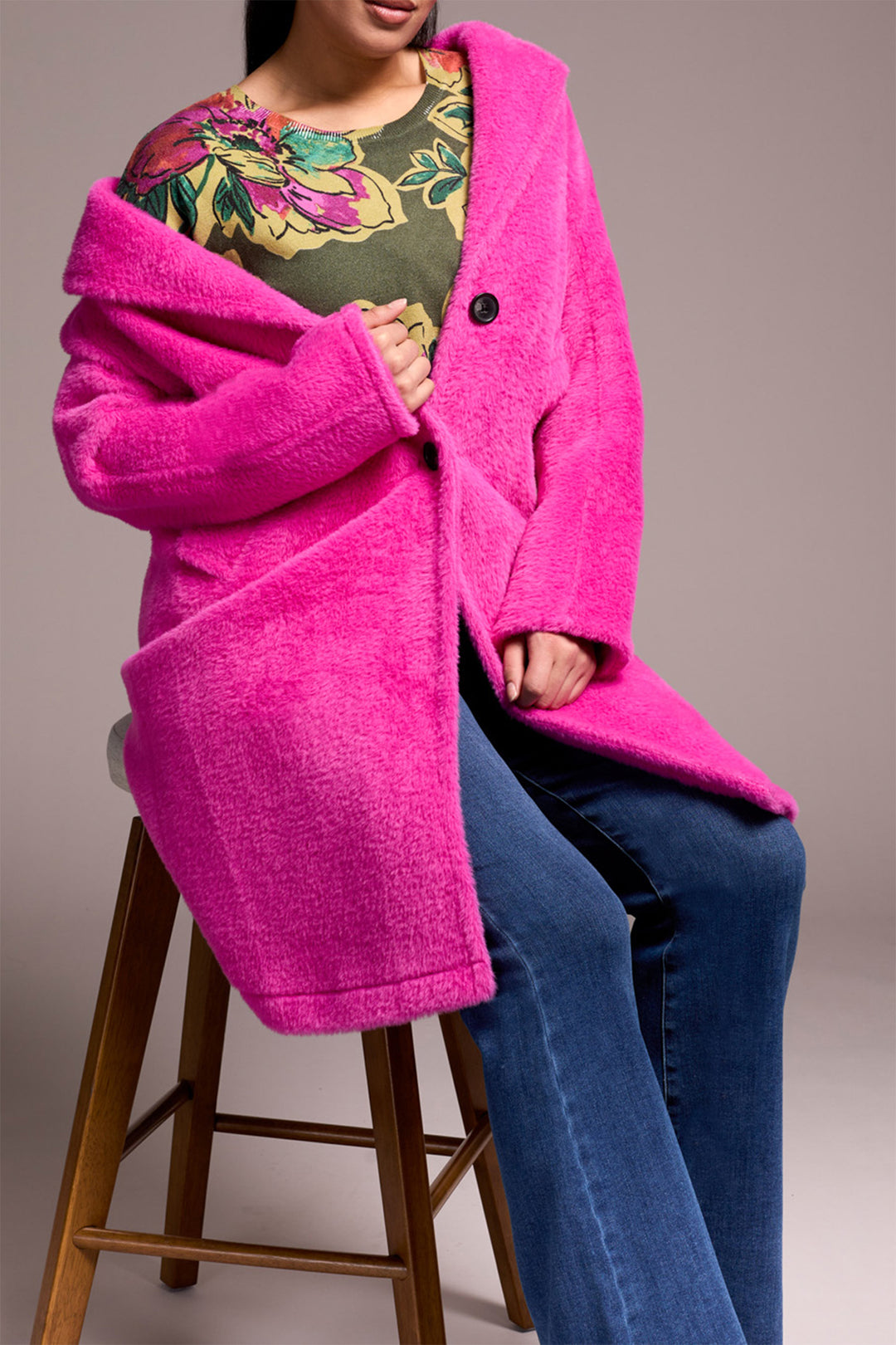 Person wearing a bright pink coat sitting on a wooden stool against a neutral background