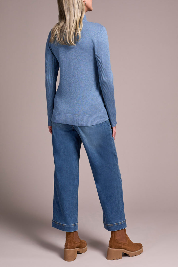 Person wearing a blue sweater and jeans with brown boots on a beige background