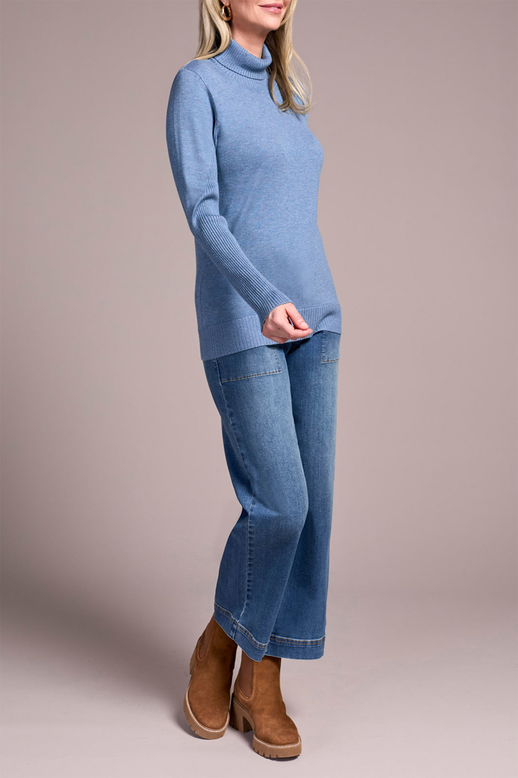 Person wearing a blue turtleneck sweater and jeans with brown boots on a plain background