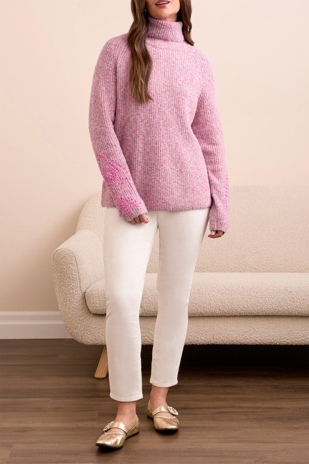 Person wearing a pink sweater and white pants standing in a room with a beige couch.