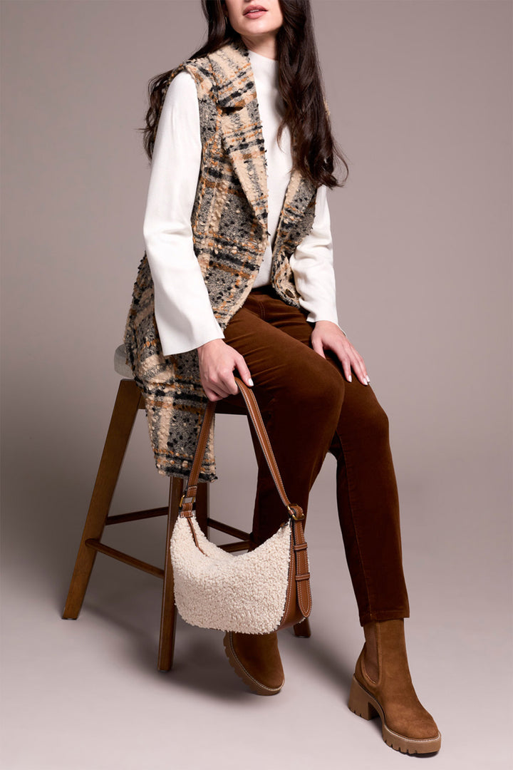 Woman sitting on a stool wearing a patterned vest, white shirt, brown pants, and brown boots with a beige handbag.