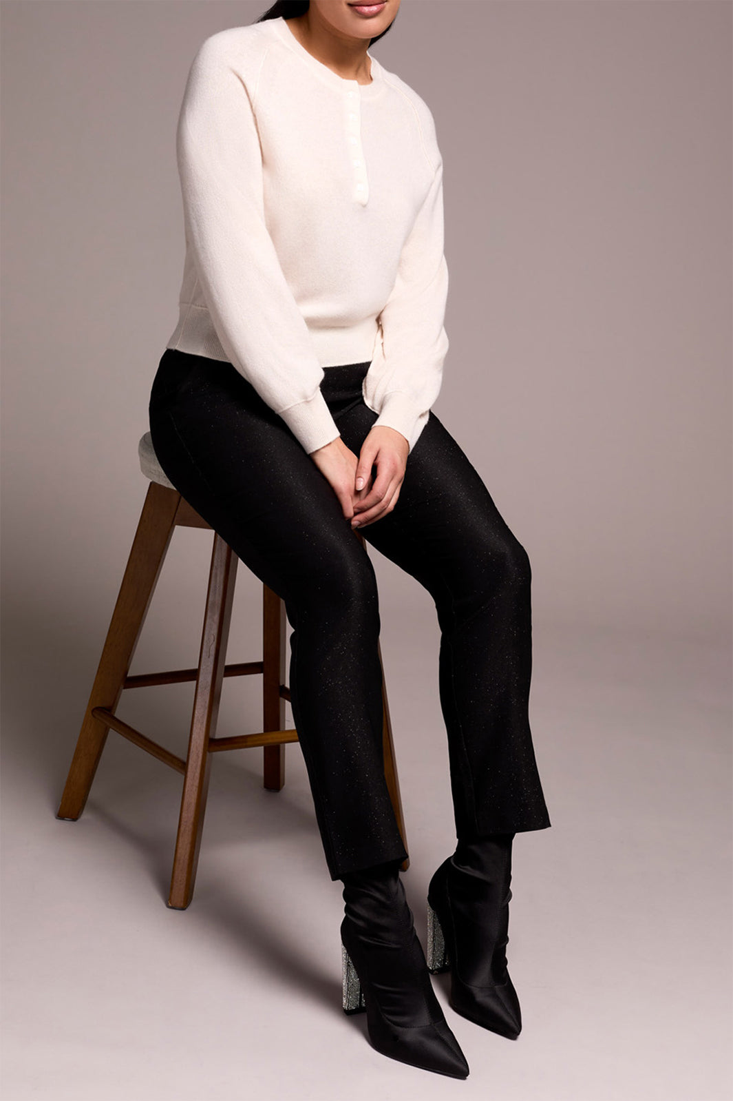 Person wearing a white sweater and black pants sitting on a wooden stool against a plain background