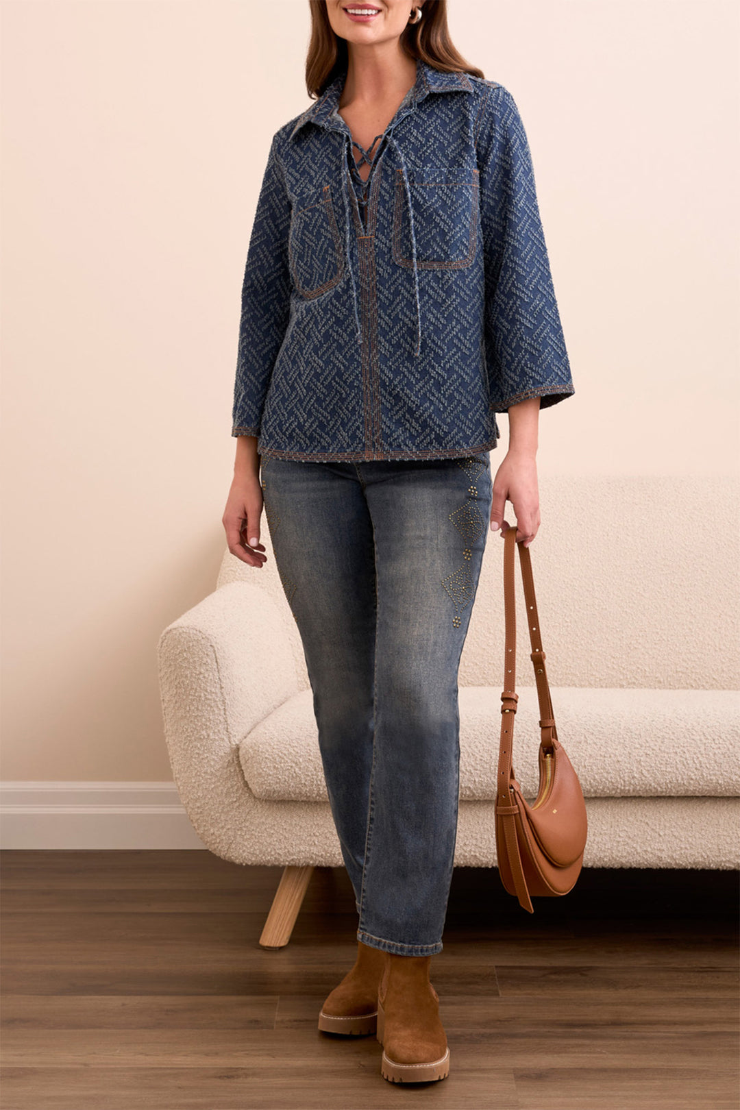 Woman wearing a blue patterned jacket, jeans, and brown boots holding a tan handbag indoors.