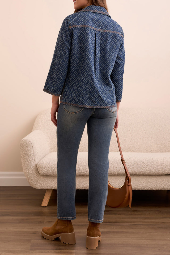 Person wearing a blue textured shirt, jeans, and brown boots standing in a room with a beige sofa.