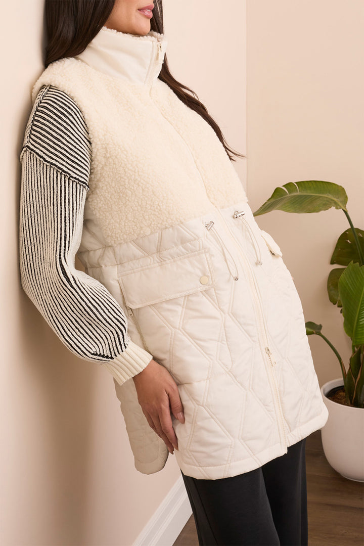 Person wearing a white puffer vest over a striped shirt against a beige wall.