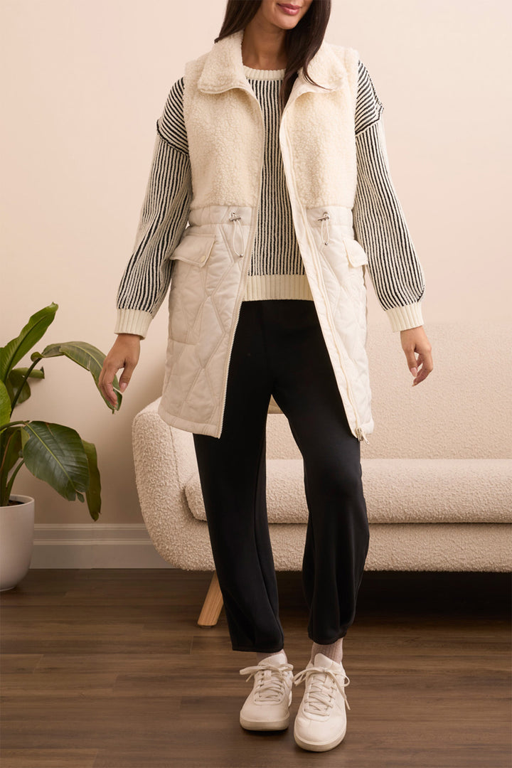 Person wearing a long cream vest over a striped sweater, black pants, and white sneakers in a living room setting.