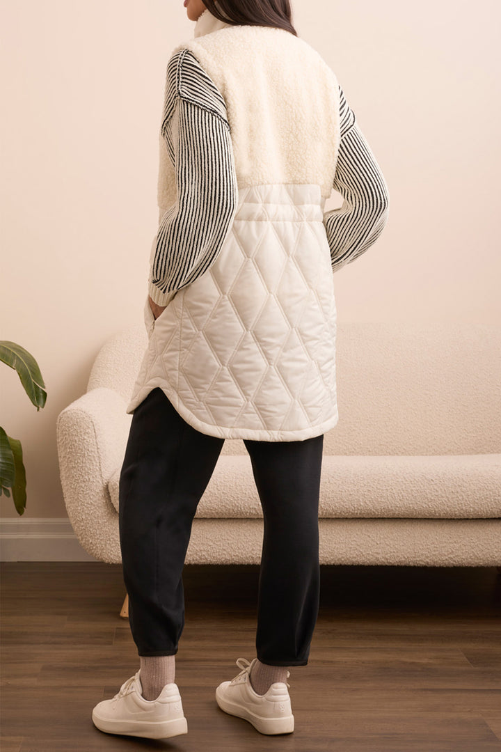 Person wearing a white quilted jacket with a striped shirt underneath, standing in a room with a beige sofa and wooden floor.
