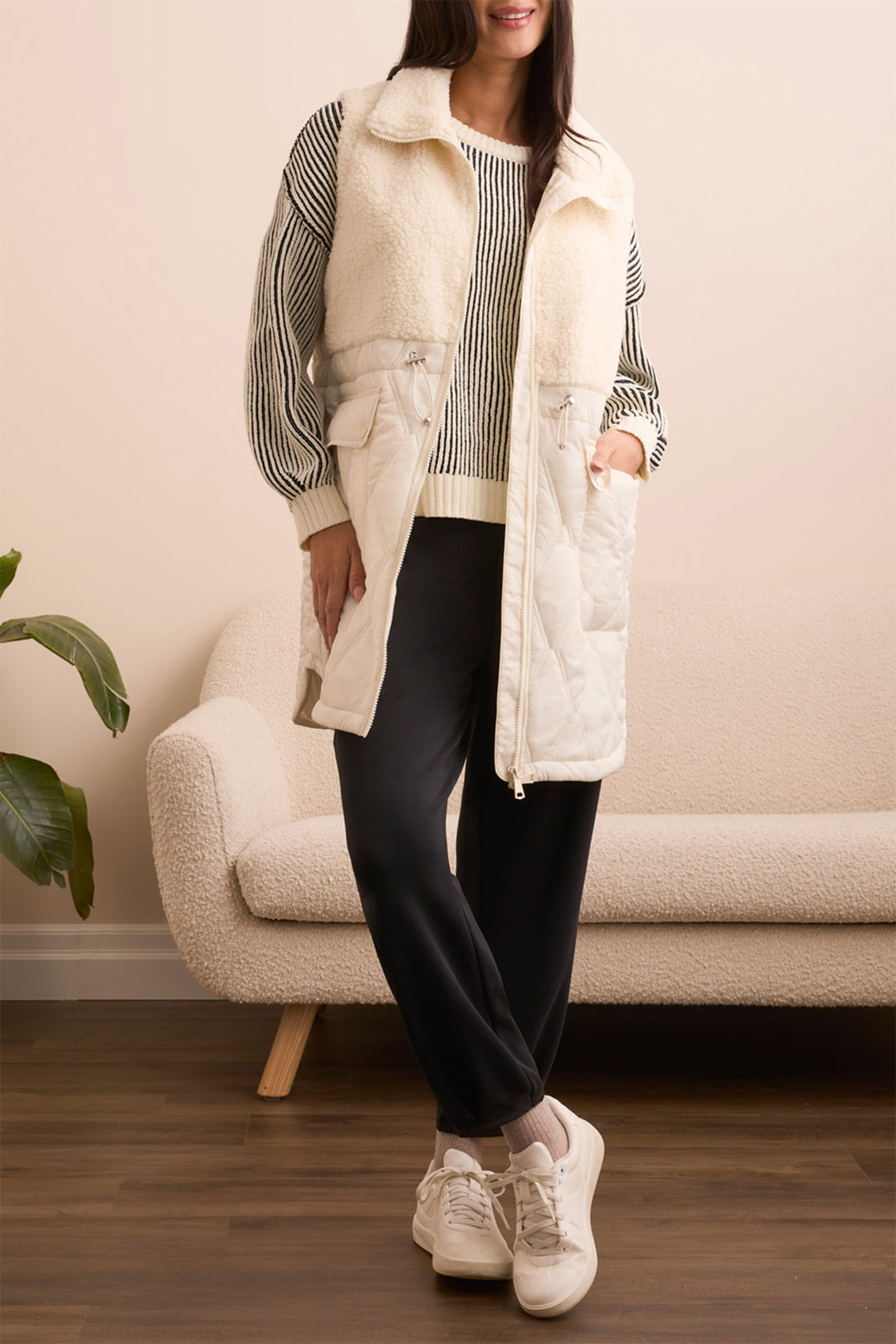 Person wearing a white puffer vest over a striped sweater with black pants and white sneakers, standing in a room with a beige sofa and plant.
