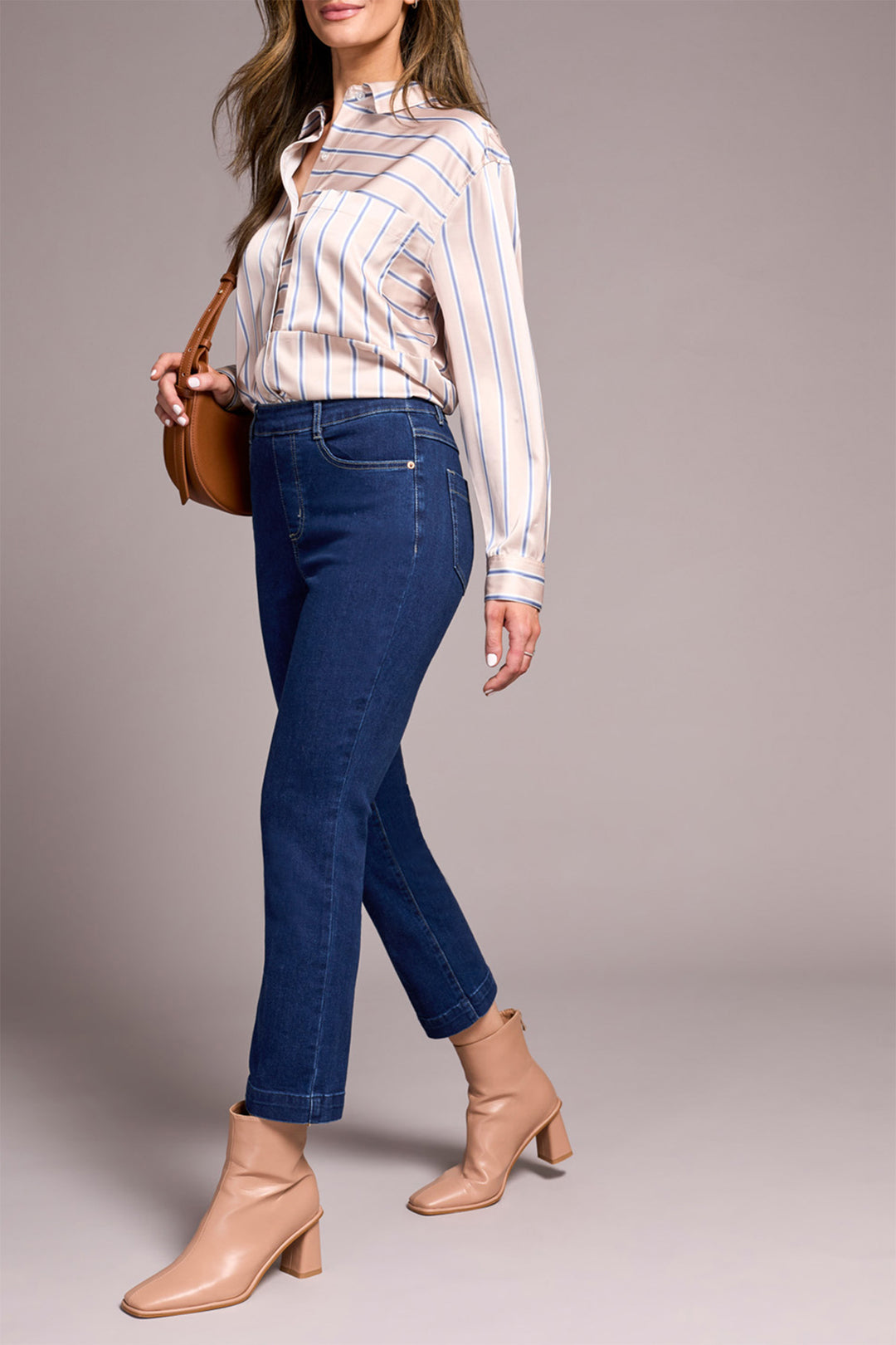 Woman wearing a striped shirt, blue jeans, and beige boots with a brown handbag on a gray background