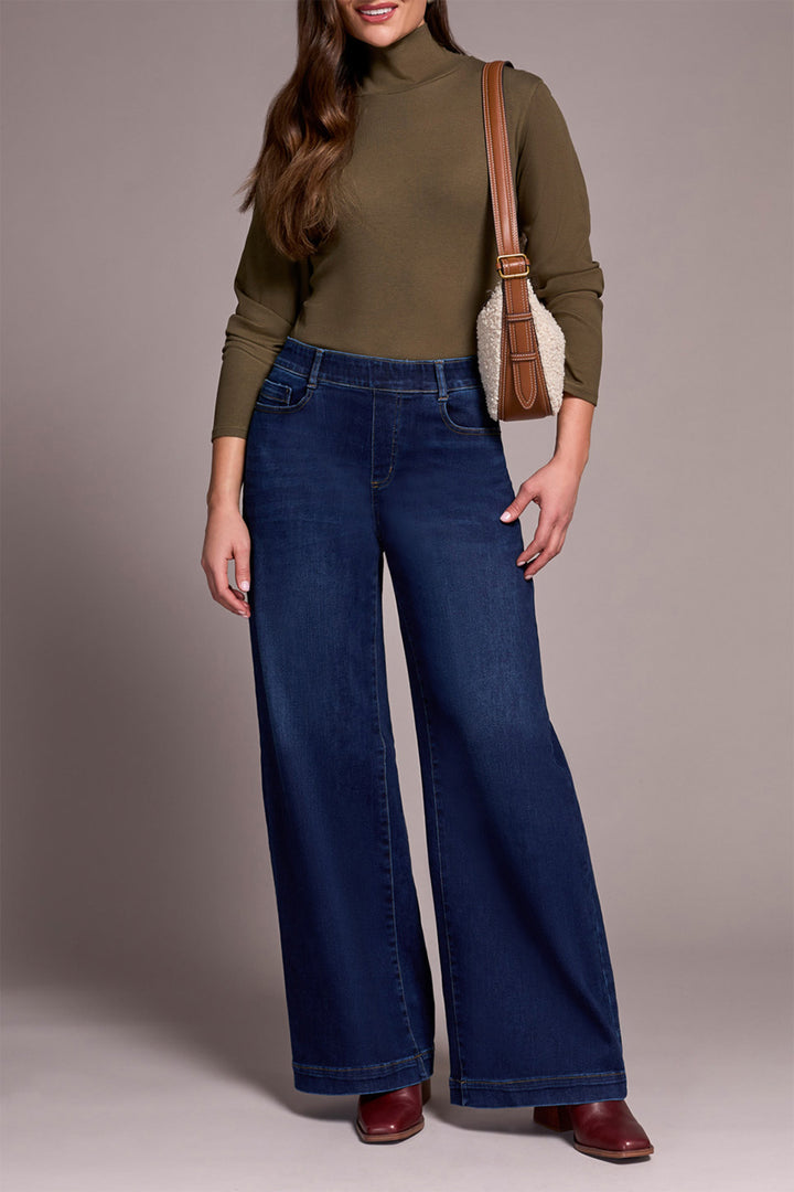 Woman wearing a green turtleneck and blue jeans with a brown bag on a neutral background