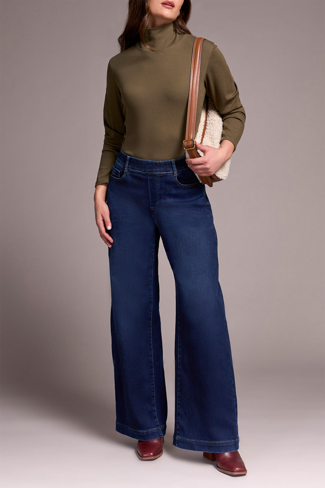 Woman wearing a green turtleneck and blue jeans, holding a beige bag against a neutral background