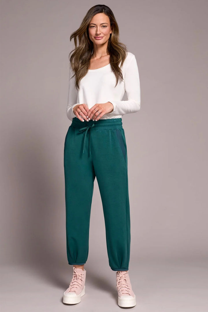Woman wearing a white top and teal pants on a plain background