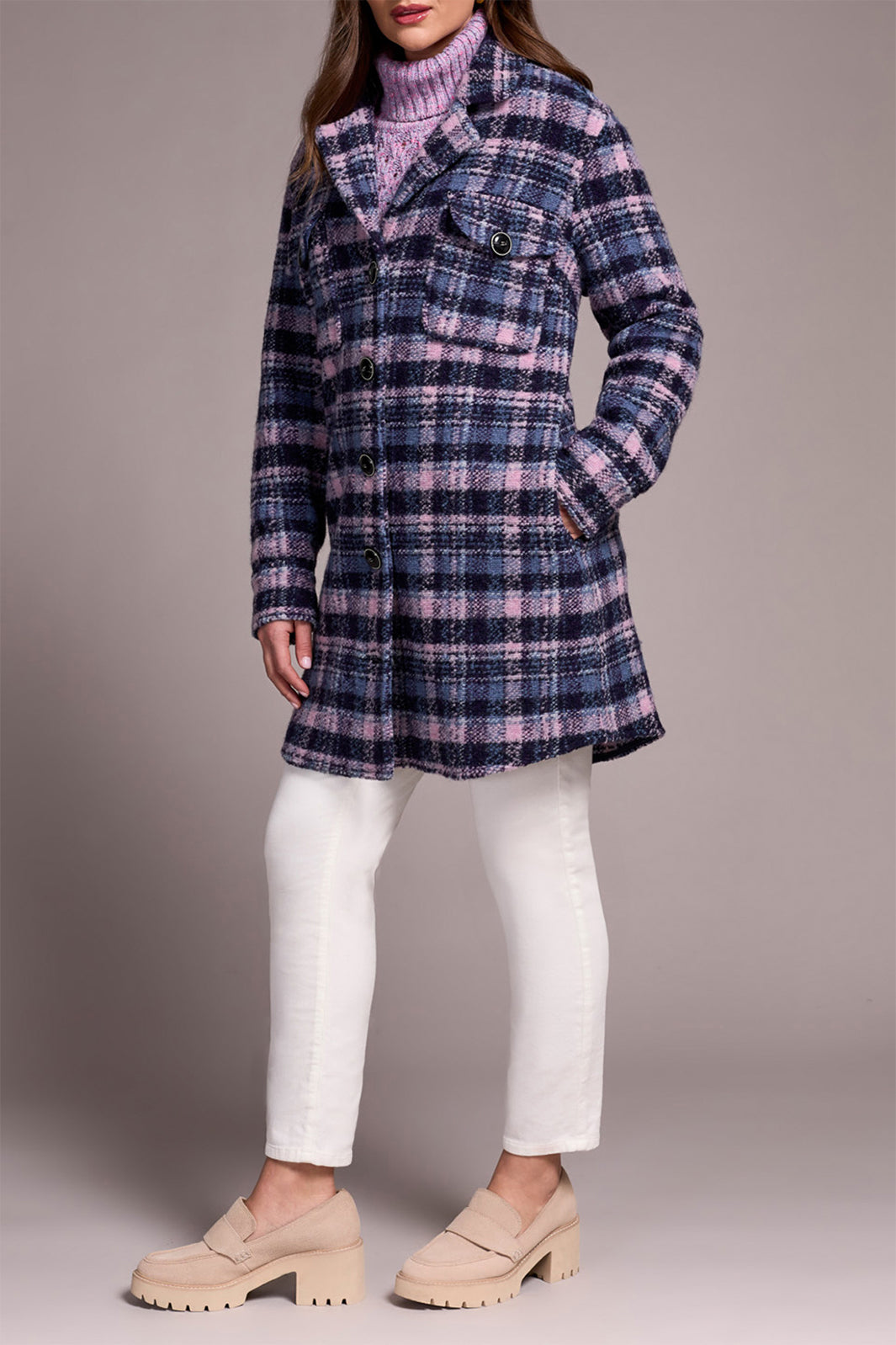 Person wearing a plaid coat over a turtleneck with white pants on a gray background
