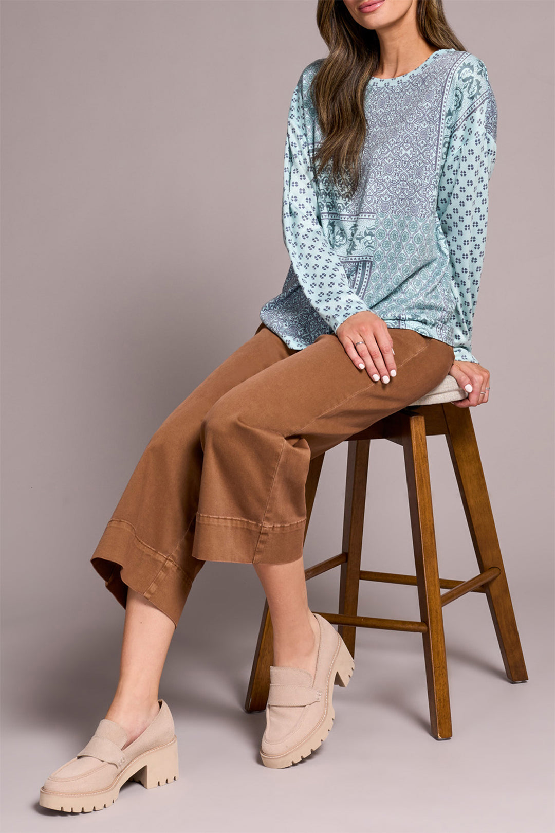 Woman sitting on a wooden stool wearing a light blue patterned top and brown pants.