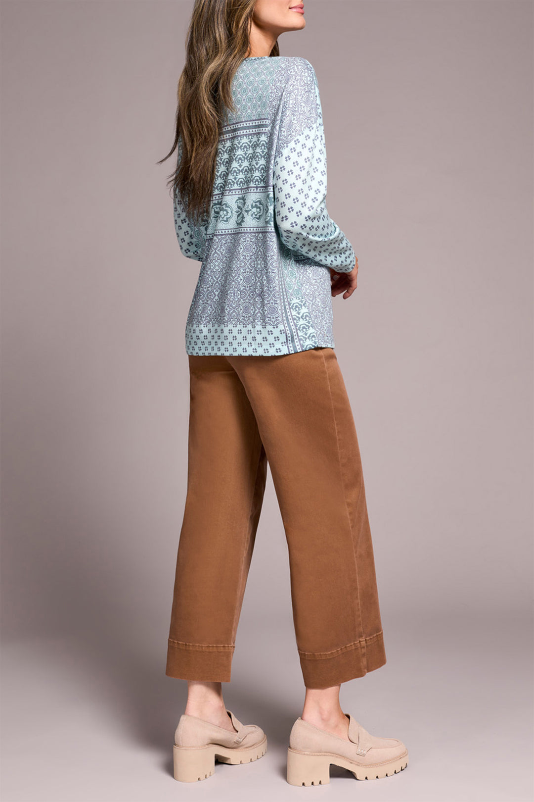 Woman wearing a light blue patterned top and brown pants on a plain background