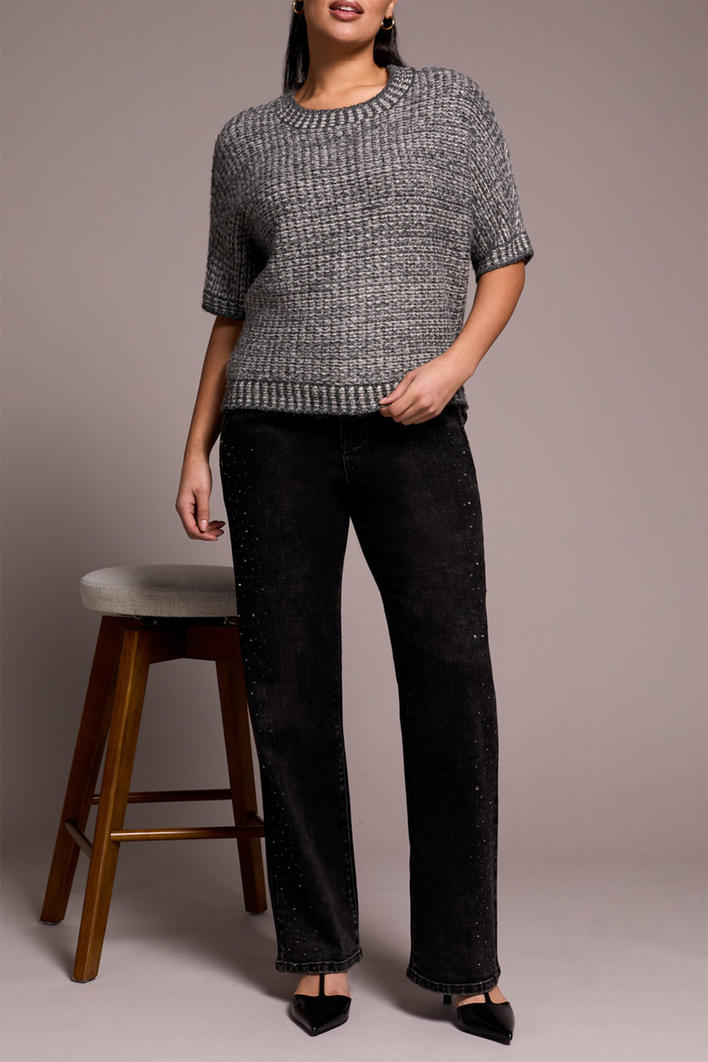 Woman wearing a gray textured sweater and black pants standing next to a wooden stool against a plain background