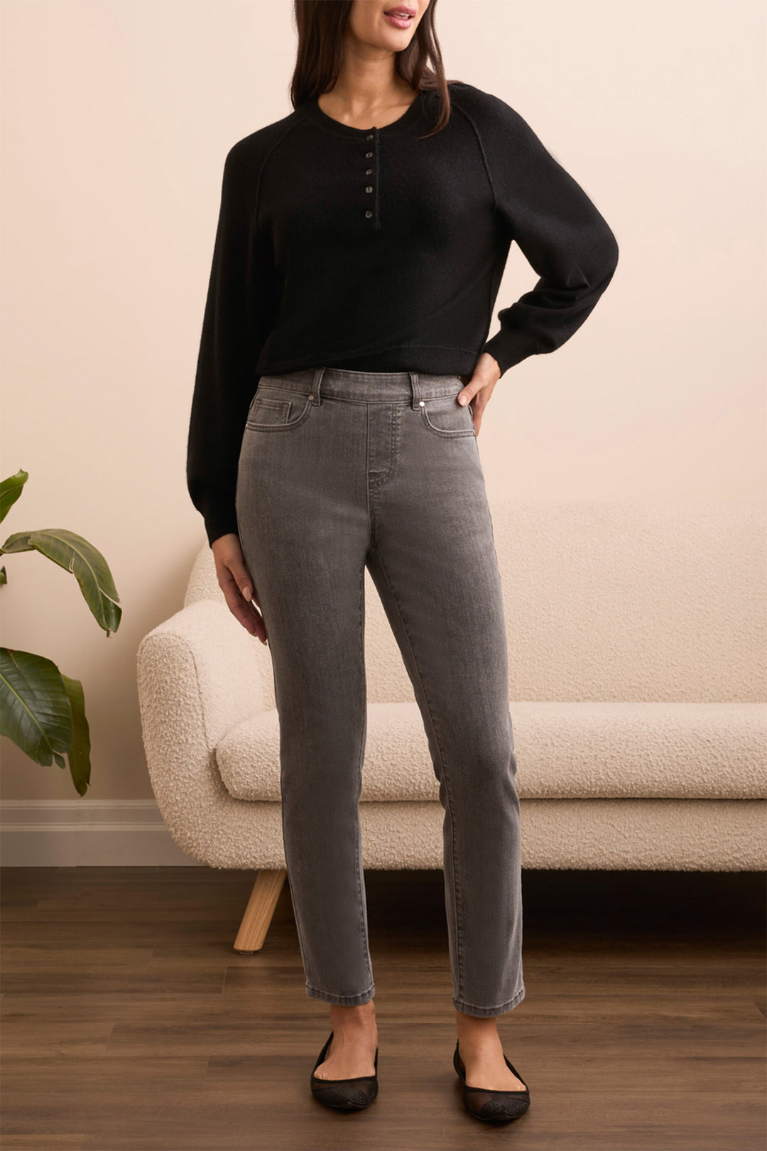 Woman wearing a black top and gray jeans standing in a room with a beige sofa and plant.