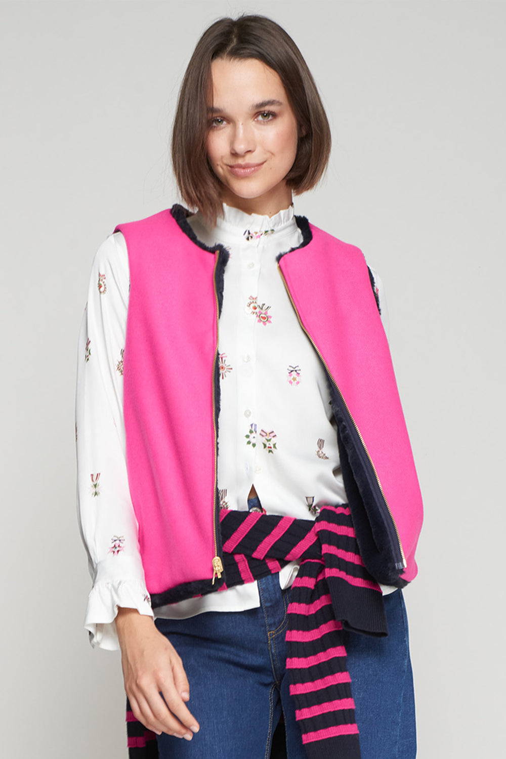 Woman wearing a pink vest over a floral blouse with a striped scarf on a gray background