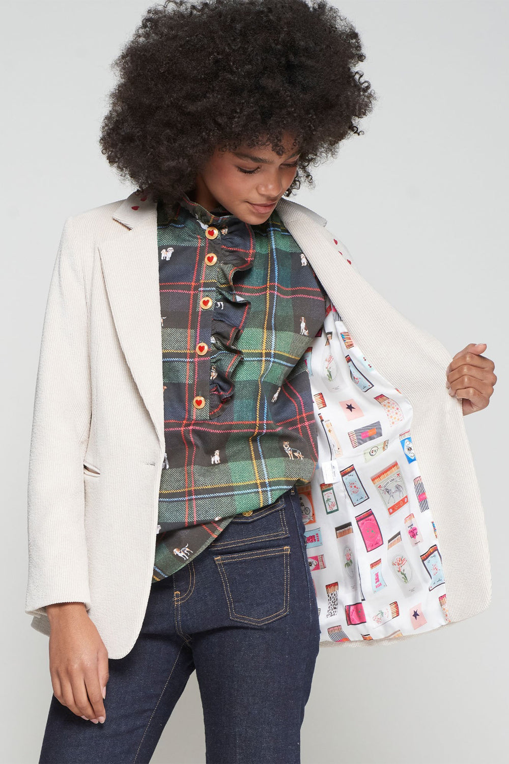 Person wearing a plaid shirt, white blazer, and jeans, holding a patterned bag against a plain background